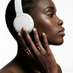 Side profile of a Black woman in white over-ear headphones, holding her ear with a hand stacked in twisted rope open rings in silver and gold tones with baguette zircon stones, on a clean white background